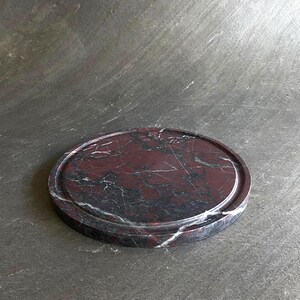 Puede incluir: Una bandeja redonda de m&aacute;rmol rojo oscuro con un borde elevado. La bandeja tiene un patr&oacute;n natural veteado.