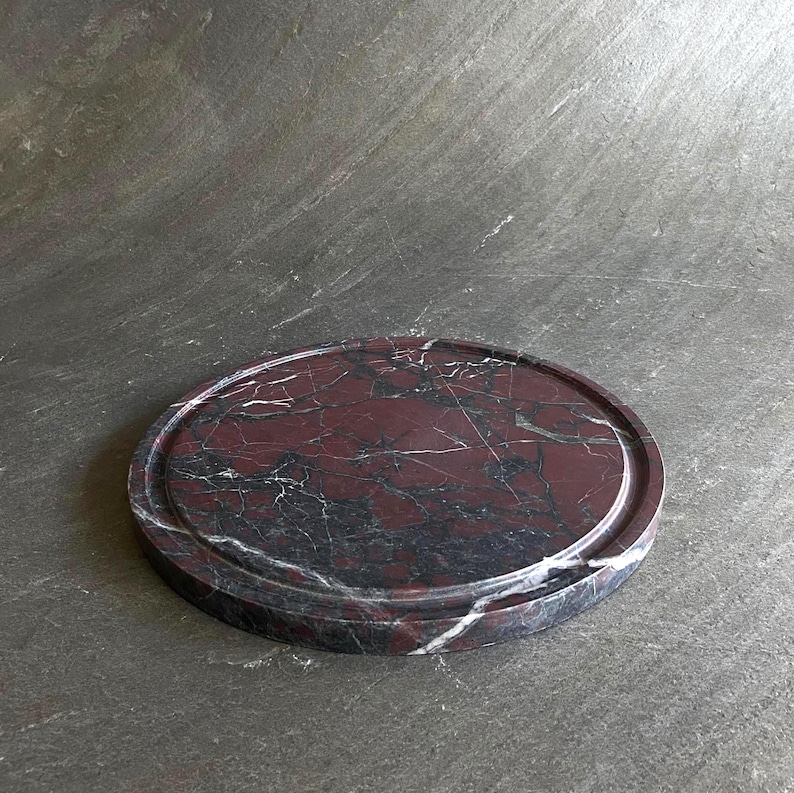 Puede incluir: Una bandeja redonda de m&aacute;rmol rojo oscuro con un borde elevado. La bandeja tiene un patr&oacute;n natural veteado.