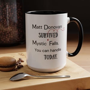 May include: White ceramic mug with a black rim and handle. The mug has black text that reads "Matt Donovan survived Mystic Falls. You can handle today."