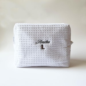 May include: White waffle-textured cosmetic bag with a zipper closure. The bag features the embroidered name "Amelia" above a small brown boot design. The bag is rectangular and appears to be made of a soft, absorbent material.