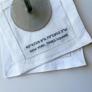 May include: White linen napkins with a blue embroidered design. The design includes the coordinates 40°4528 8"N 73°59'05 5"W and the text "NEW YORK, TIMES SQUARE".