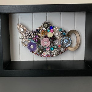 May include: A framed artwork featuring a teapot design. The teapot is crafted from an assortment of colorful jewels, beads, and decorative elements. The frame is black, and the background is white with vertical lines. The teapot handle is gold.