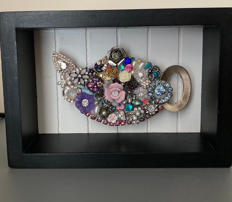 May include: A framed artwork featuring a teapot design. The teapot is crafted from an assortment of colorful jewels, beads, and decorative elements. The frame is black, and the background is white with vertical lines. The teapot handle is gold.