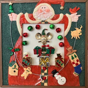 May include: A Christmas-themed wall hanging featuring a Santa Claus figure with a red coat and white trim. The artwork includes various festive elements like bells, gifts, a snowman, and a reindeer. The background is green with a brown frame.