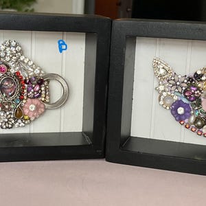 May include: Two framed artworks each feature a teapot design crafted from an assortment of sparkling jewels and embellishments. The teapots are set against a white background with vertical lines, framed in black. One teapot has a blue gem.