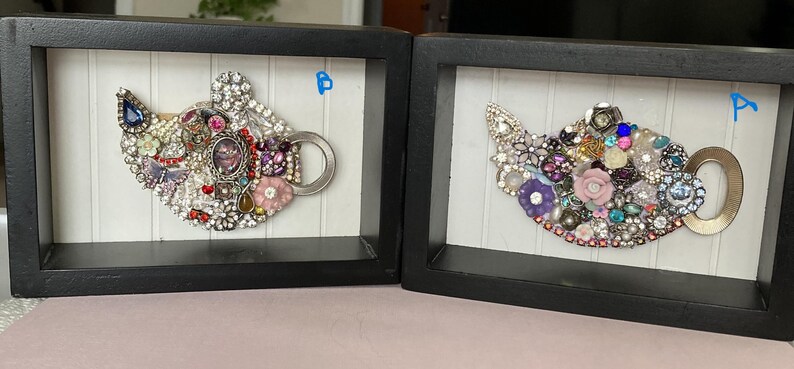 May include: Two framed artworks each feature a teapot design crafted from an assortment of sparkling jewels and embellishments. The teapots are set against a white background with vertical lines, framed in black. One teapot has a blue gem.