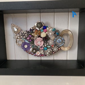 May include: A framed artwork featuring a teapot design. The teapot is adorned with an assortment of colorful jewels, beads, and decorative elements. The handle is gold-colored, and the background is white with vertical lines.