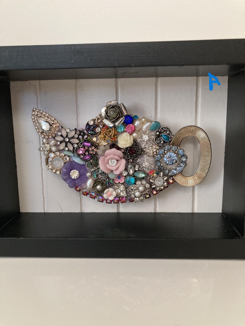 May include: A framed artwork featuring a teapot design. The teapot is adorned with an assortment of colorful jewels, beads, and decorative elements. The handle is gold-colored, and the background is white with vertical lines.