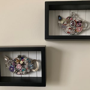 May include: Two decorative teapot wall art pieces. Each teapot is adorned with an assortment of colorful jewels, beads, and faux pearls. The teapots are set within black shadow boxes with a white, vertical-slat background.