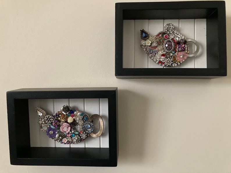 May include: Two decorative teapot wall art pieces. Each teapot is adorned with an assortment of colorful jewels, beads, and faux pearls. The teapots are set within black shadow boxes with a white, vertical-slat background.