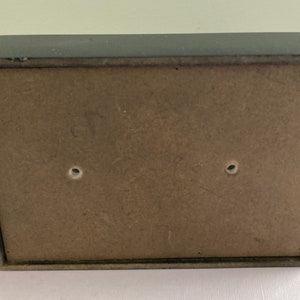 May include: A small, rectangular, black wooden box with a light brown back panel. The back panel has two holes. The box appears to be empty. The box is likely used for storage or display.