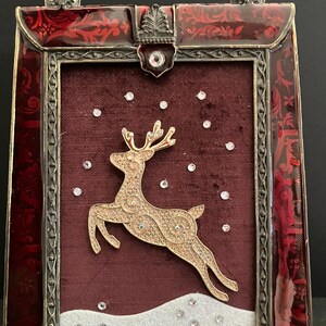 May include: A decorative Christmas-themed picture frame with a red and gold color scheme. The frame features a gold reindeer leaping across a burgundy background with faux snow and sparkling accents. The frame has a handle.