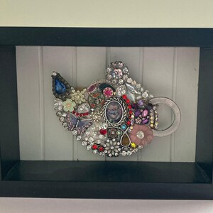 May include: A framed artwork featuring a jeweled cat head design. The cat head is composed of various colorful jewels, including rhinestones, beads, and vintage jewelry pieces. The frame is black.