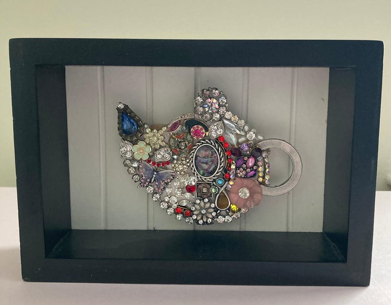 May include: A framed artwork featuring a jeweled cat head design. The cat head is composed of various colorful jewels, including rhinestones, beads, and vintage jewelry pieces. The frame is black.