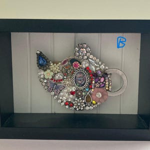 May include: A framed artwork featuring a teapot design, embellished with an assortment of colorful jewels and rhinestones. The teapot is set against a white background and enclosed in a black frame. The artwork includes a variety of shapes and colors.