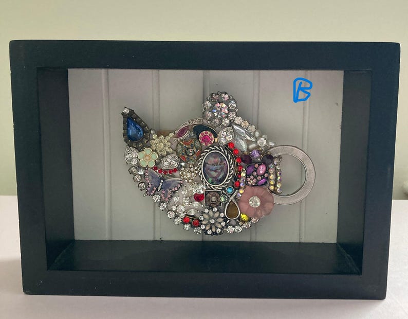 May include: A framed artwork featuring a teapot design, embellished with an assortment of colorful jewels and rhinestones. The teapot is set against a white background and enclosed in a black frame. The artwork includes a variety of shapes and colors.