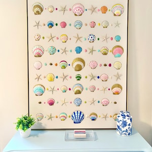 May include: A framed artwork featuring a collection of colorful seashells and starfish. The shells are painted in various hues, including pink, yellow, and blue, and are arranged on a light background. The artwork is displayed above a light blue dresser.