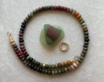 May include: A circular beaded necklace featuring small, smooth, multi-colored stone beads in shades of green, brown, red, and tan. The necklace includes a gold-toned metal clasp and is arranged around a central green and brown sea glass piece.