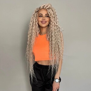 May include: A woman with long, blonde, curly hair is wearing a bright orange crop top and black pants. She is smiling and looking to the side.