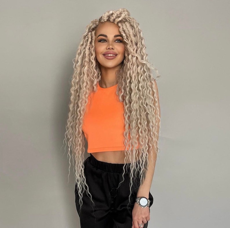 May include: A woman with long, blonde, curly hair is wearing a bright orange crop top and black pants. She is smiling and looking to the side.