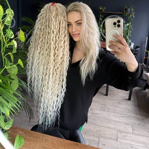 May include: A woman with long, wavy, blonde hair is holding a phone in front of her face. She is wearing a black shirt and has a strand of hair tied with a red hair tie.