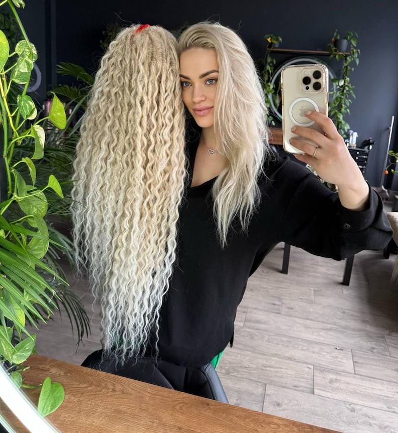 May include: A woman with long, wavy, blonde hair is holding a phone in front of her face. She is wearing a black shirt and has a strand of hair tied with a red hair tie.