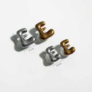 May include: Four letter "E" stud earrings. Two silver glitter and two gold glitter earrings are displayed. The silver earrings measure 21mm x 17mm and 17mm x 12mm. The gold earrings are 17mm x 17mm and 17mm x 12mm.