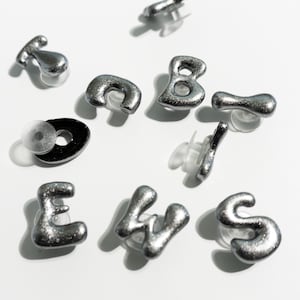 May include: Silver-colored stud earrings spelling out "EWS" and a letter "B", plus a black and silver earring. The earrings have a textured, metallic finish and are set against a white background. The earrings are small and likely for pierced ears.