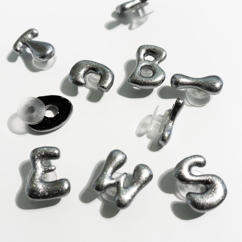 May include: Silver-colored stud earrings spelling out "EWS" and a letter "B", plus a black and silver earring. The earrings have a textured, metallic finish and are set against a white background. The earrings are small and likely for pierced ears.