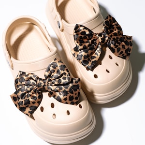 Può includere: Un paio di zoccoli beige con fiocchi con stampa leopardata nera e marrone. I fiocchi sono in materiale lucido. Gli zoccoli hanno diversi fori e un cinturino sul retro. Scarpe di colore neutro.