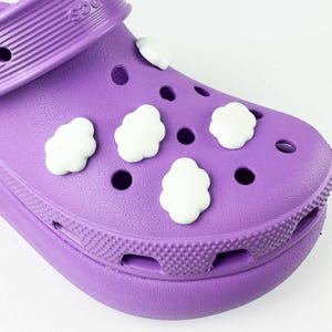 May include: Close-up of a purple clog shoe with white cloud-shaped charms. The shoe has multiple ventilation holes and a textured surface. The charms are evenly spaced across the top of the shoe.