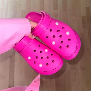 Puede incluir: Un par de zuecos de color rosa brillante con adornos de perlas blancas. Los zuecos tienen un diseño clásico con múltiples agujeros en toda la superficie.