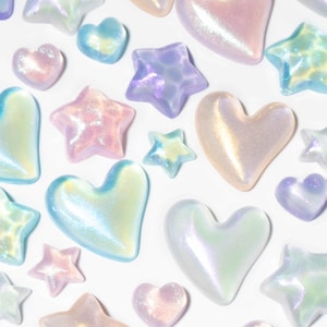 May include: A collection of iridescent heart and star-shaped embellishments in pastel shades. The shapes are scattered on a white background. The colours include pink, blue, purple, and yellow, with a shimmering, pearlescent finish. These are ideal for crafts.