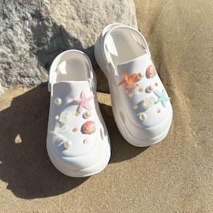 Könnte beinhalten: Weiße Plateau-Clogs, verziert mit Muscheln, Seesternen und Perlen. Die Clogs haben einen lässigen, sommerlichen Stil.
