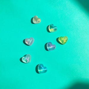 Op de afbeelding: Zeven hartvormige, doorschijnende harsstukken in tinten groen, blauw en iriserend wit zijn verspreid op een turquoise achtergrond. De harten hebben een glinsterend, glimmend effect, dat het licht vangt. De afbeelding is helder verlicht met schaduwen.