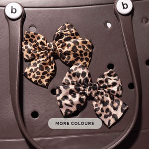 Könnte beinhalten: Drei Schleifen mit Leopardenmuster auf dunklem braunen Hintergrund. Die Schleifen bestehen aus einem seidigen Material mit einem beige-schwarzen Muster. Der Text "MORE COLOURS" wird auf einem weißen Rechteck angezeigt.