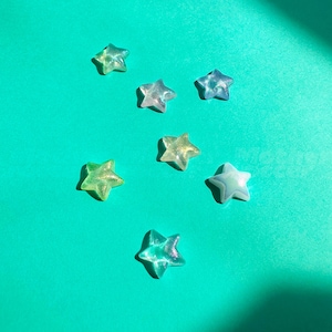 Op de afbeelding: Zeven doorschijnende stervormige kralen in tinten groen, blauw en geel zijn verspreid op een turquoise achtergrond. De kralen hebben een glinsterende, iriserende kwaliteit die het licht vangt. De sterren vari&euml;ren enigszins in grootte en kleur.