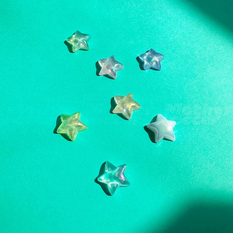 Op de afbeelding: Zeven doorschijnende stervormige kralen in tinten groen, blauw en geel zijn verspreid op een turquoise achtergrond. De kralen hebben een glinsterende, iriserende kwaliteit die het licht vangt. De sterren vari&euml;ren enigszins in grootte en kleur.