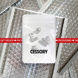 May include: A clear plastic bag containing silver-colored heart, star, and letter-shaped accessories. The bag has the word "CESSORY" printed on it. The background is a silver bubble wrap material.