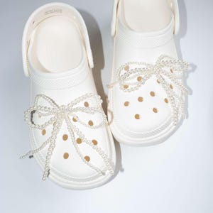 Peut inclure: Sabots blancs avec un détail de nœud en perles. Les sabots ont une silhouette classique et sont décorés d'un nœud en perles à l'avant.