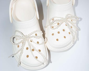 May include: A pair of white clogs with pearl embellishments. The clogs have a classic design with multiple ventilation holes and a strap. Each clog is adorned with a pearl bow, adding a touch of elegance to the casual footwear.