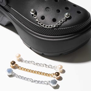 May include: A black platform clog shoe featuring a silver-toned metal chain charm attached to its surface. Below the shoe, three additional decorative chain charms are displayed: one with white pearl-like beads, one gold-toned, and one silver-toned.