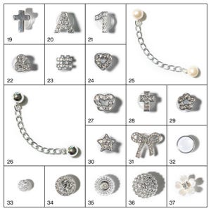 May include: A collection of silver-toned charms and embellishments, including crosses, letters, numbers, hearts, stars, bows, and floral designs. Some charms feature clear crystal accents and chain details. The items are arranged in a grid.