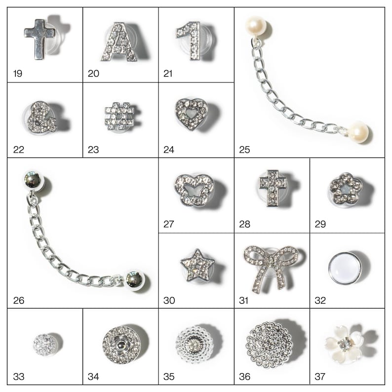 May include: A collection of silver-toned charms and embellishments, including crosses, letters, numbers, hearts, stars, bows, and floral designs. Some charms feature clear crystal accents and chain details. The items are arranged in a grid.