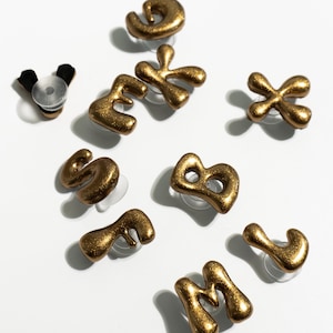 May include: Gold-colored, glittery alphabet letter stud earrings. The letters are in a unique, abstract font. The earrings are displayed on a white surface. A bottle stopper is also visible.