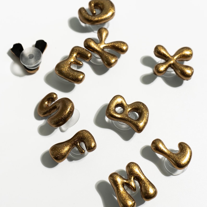 May include: Gold-colored, glittery alphabet letter stud earrings. The letters are in a unique, abstract font. The earrings are displayed on a white surface. A bottle stopper is also visible.