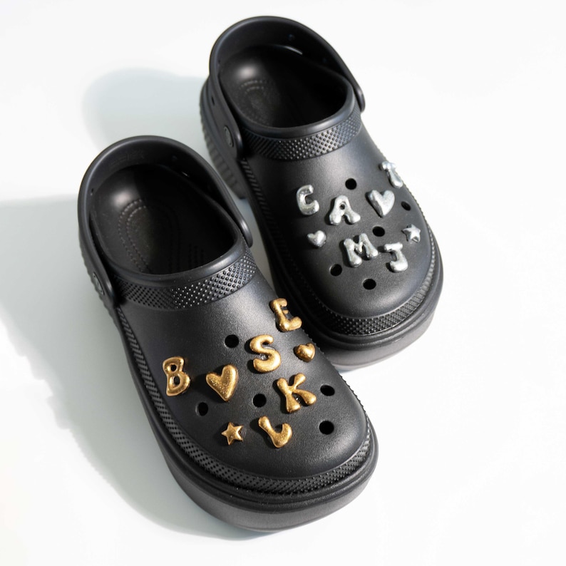 May include: Two black clogs with decorative charms. One clog has gold charms spelling out "B.E.S.K." and a star and heart. The other clog has silver charms spelling "CAV.I.MJ" with heart and star charms. The clogs have a platform sole.