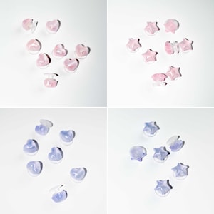 Op de afbeelding: Een verzameling hart- en stervormige oorstekers in roze en paarse tinten. De oorbellen zijn in groepen gerangschikt op een wit oppervlak. De oorbellen hebben een doorschijnend uiterlijk.