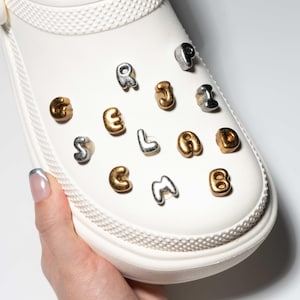 Peut inclure: Une chaussure blanche ornée de breloques lettres métalliques dorées et argentées épelant le mot "SEABIRD". La chaussure est tenue par une main avec un ongle peint.