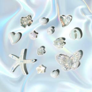 Puede incluir: Una colección de decoraciones iridiscentes para uñas en varias formas, incluyendo corazones, estrellas, un lazo, una mariposa y una estrella de mar. Las decoraciones transparentes tienen un efecto brillante, sobre un fondo azul claro iridiscente.
